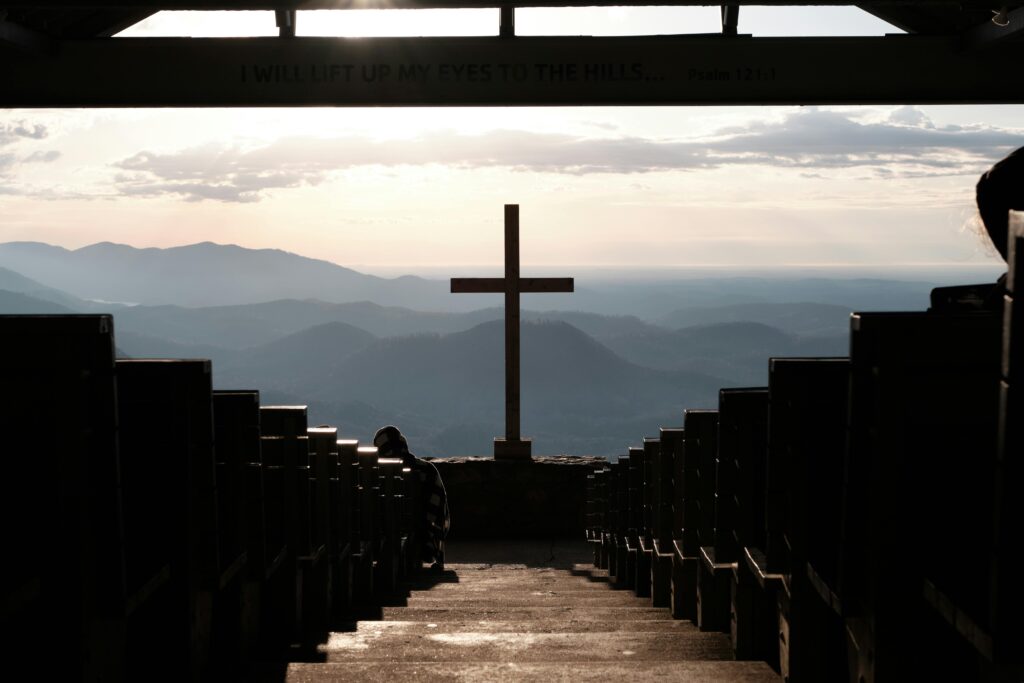 Serene view of a chapel cross overlooking mountains at sunset, with benches leading to the altar.